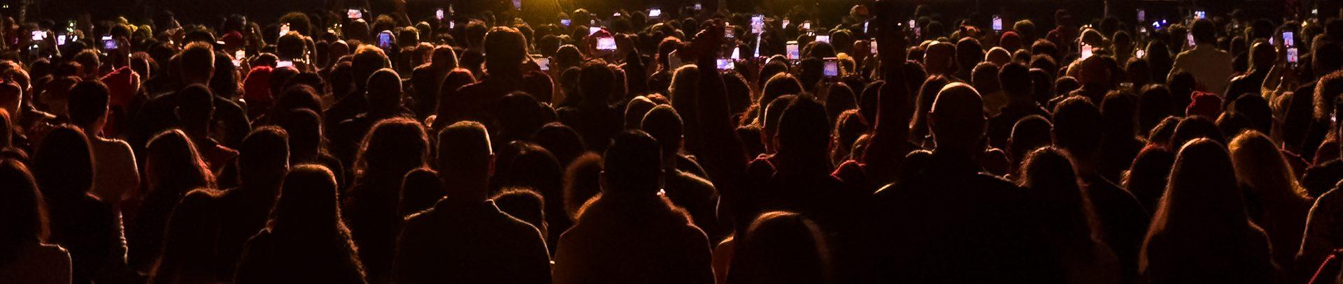 A dense concert audience seen from behind, back-lit warm amber from the stage, heads and shoulders in near-silhouette with rim light on hair, and dozens of raised phone screens glowing pale blue across the upper third.