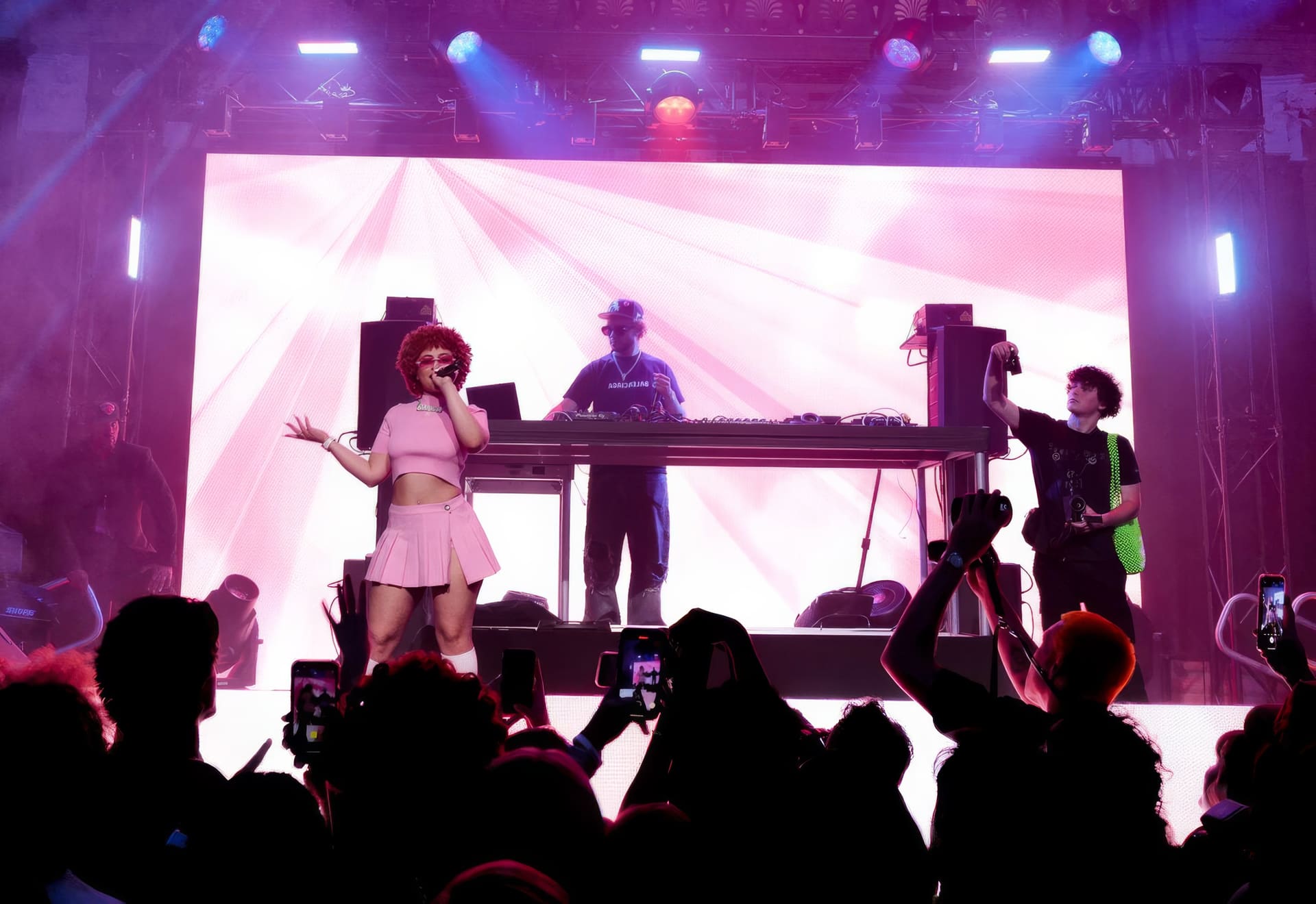 Ice Spice performing at the Bose launch — a full-width LED wall behind her throwing pink radiating rays, a DJ at the console mid-stage, and the audience in silhouette with phones raised.