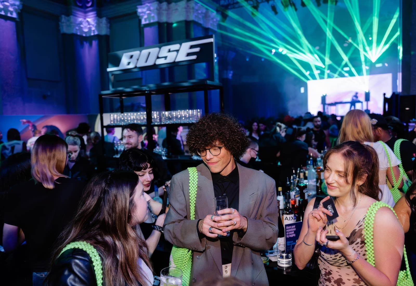 Guests with drinks in front of a lit BOSE sign at the bar, green laser fans crossing the room above them and a lit screen beyond.