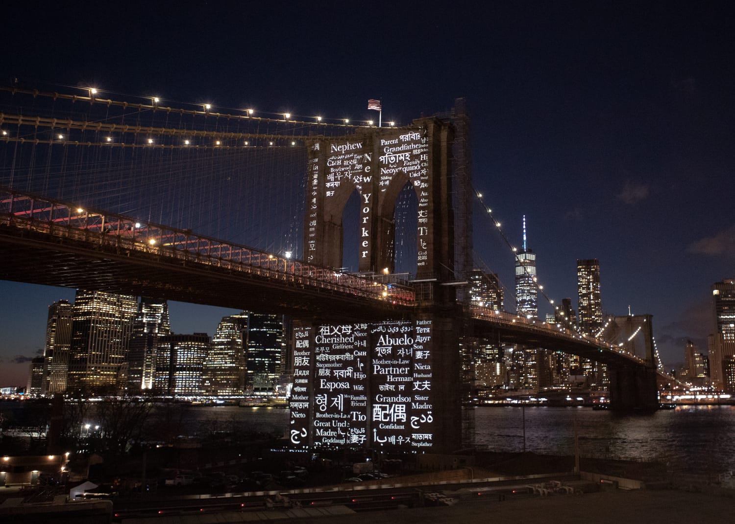 Words for family and kinship in many languages projected across the granite tower and pier of the Brooklyn Bridge at night, the Manhattan skyline beyond.