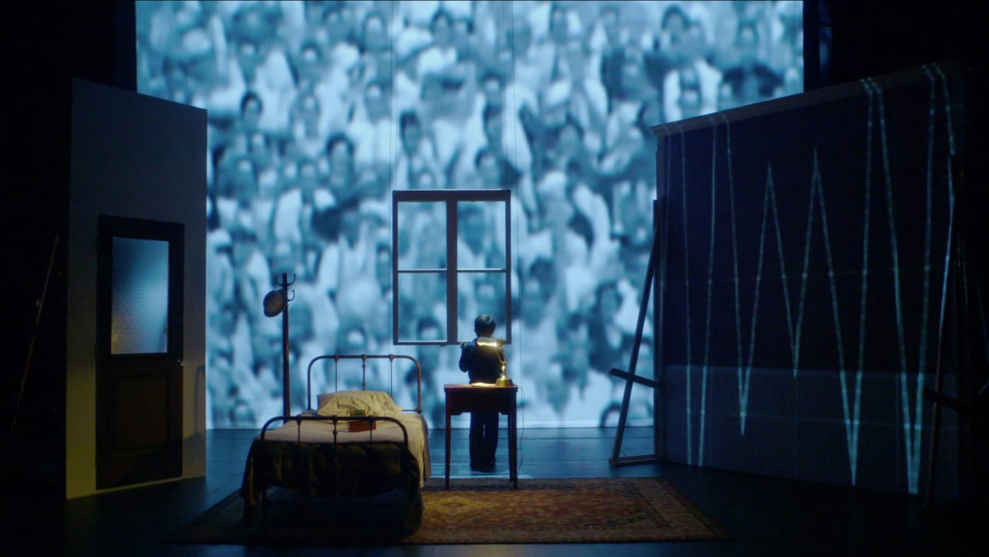 A theatrical set — a lone figure at a desk before a projected wall of faces.