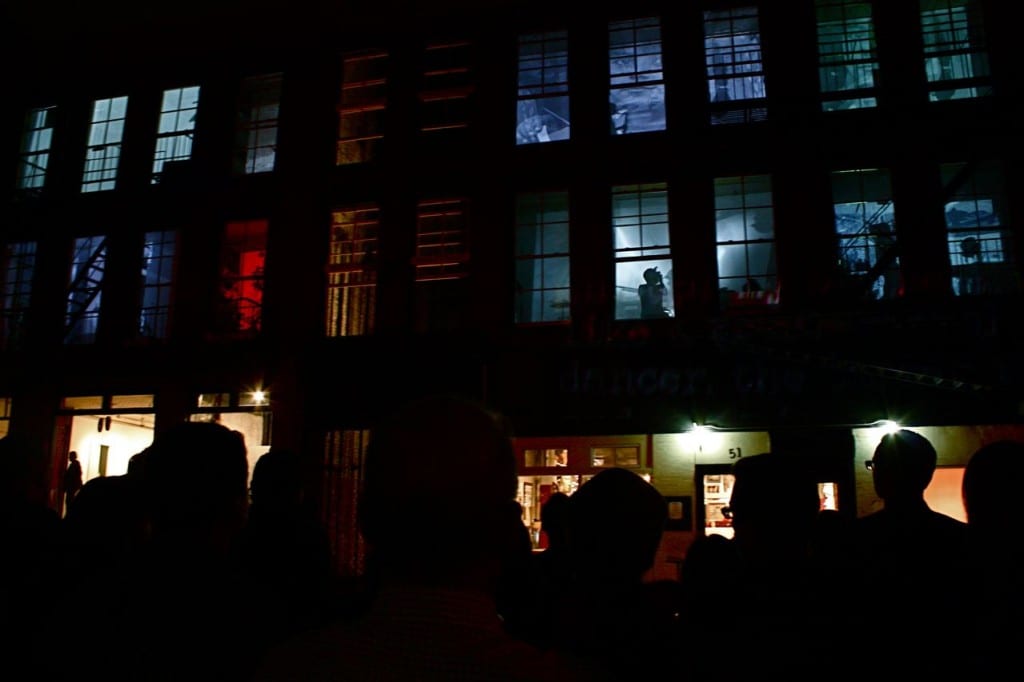 The lit windows of a dark industrial building at night, silhouettes gathered below.