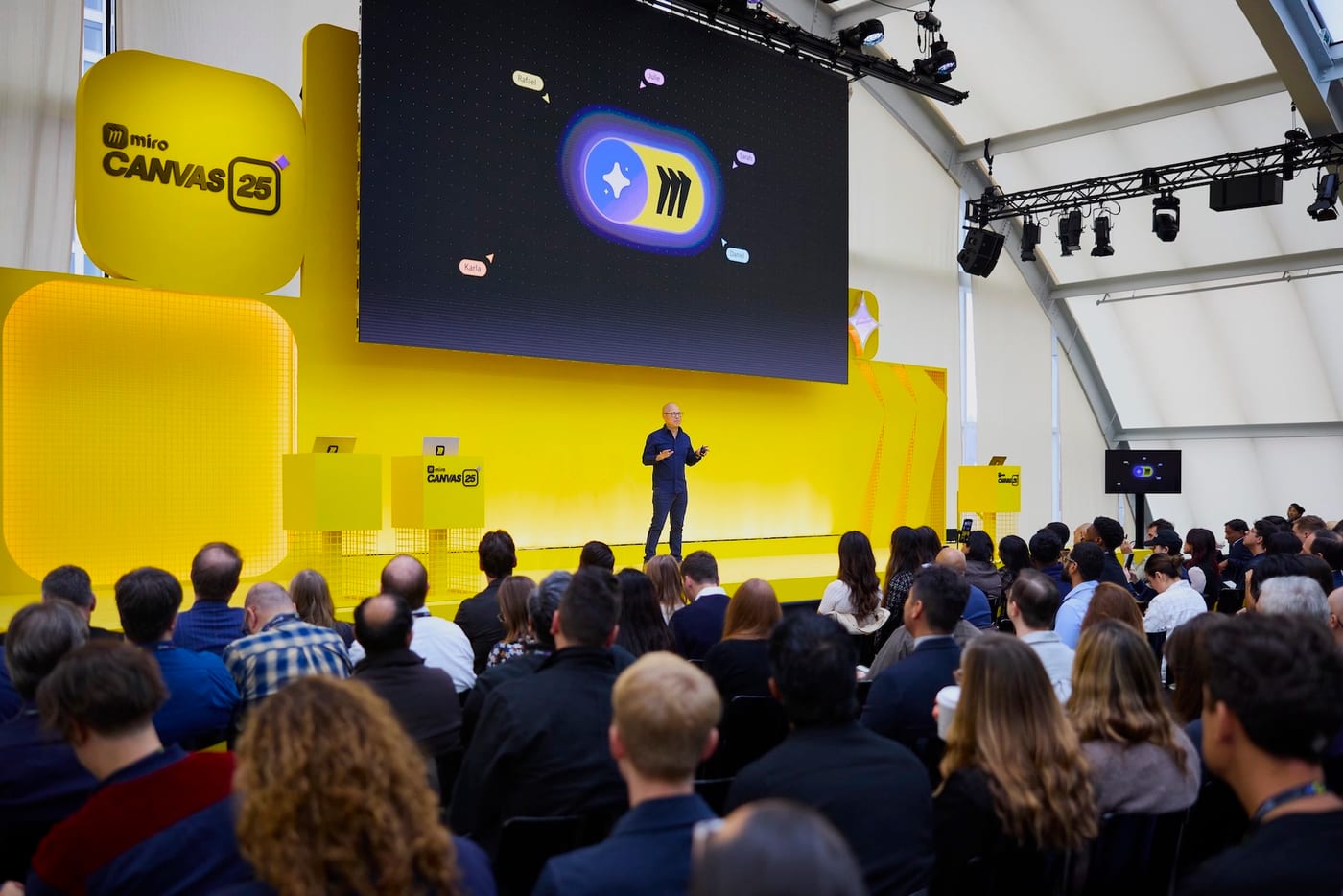 A keynote stage at Miro Canvas — a speaker before a large screen, the audience filling the room.