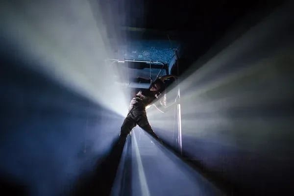 A lone figure crossing shafts of light in a dark immersive set.